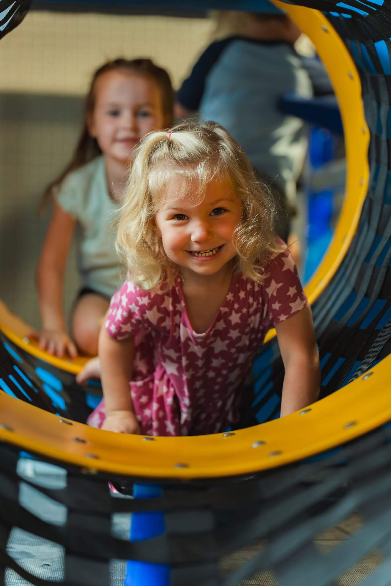 Smiling girl in a star-patterned dress crawls through a tunnel in the Climbing Zone, an indoor play area at Sunflower Discovery Center in Decorah, IA.