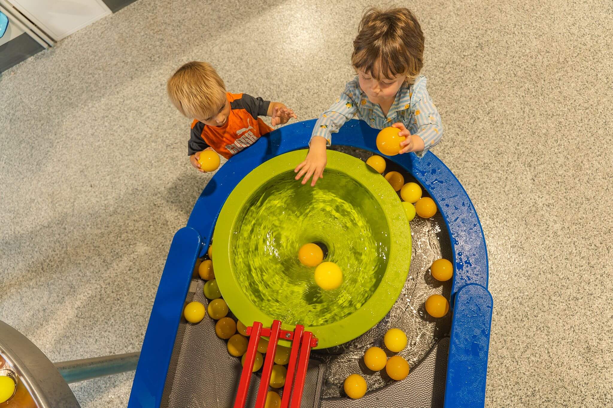 Two children enjoy family fun at a water activity table with a spinning green basin, exploring STEAM learning as they play with yellow balls.