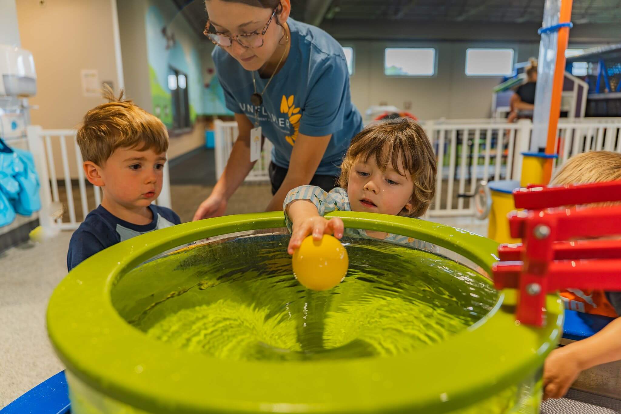 Three children play with a yellow ball in a fun water activity at the Sunflower Discovery Center’s indoor play area, supervised by an adult.